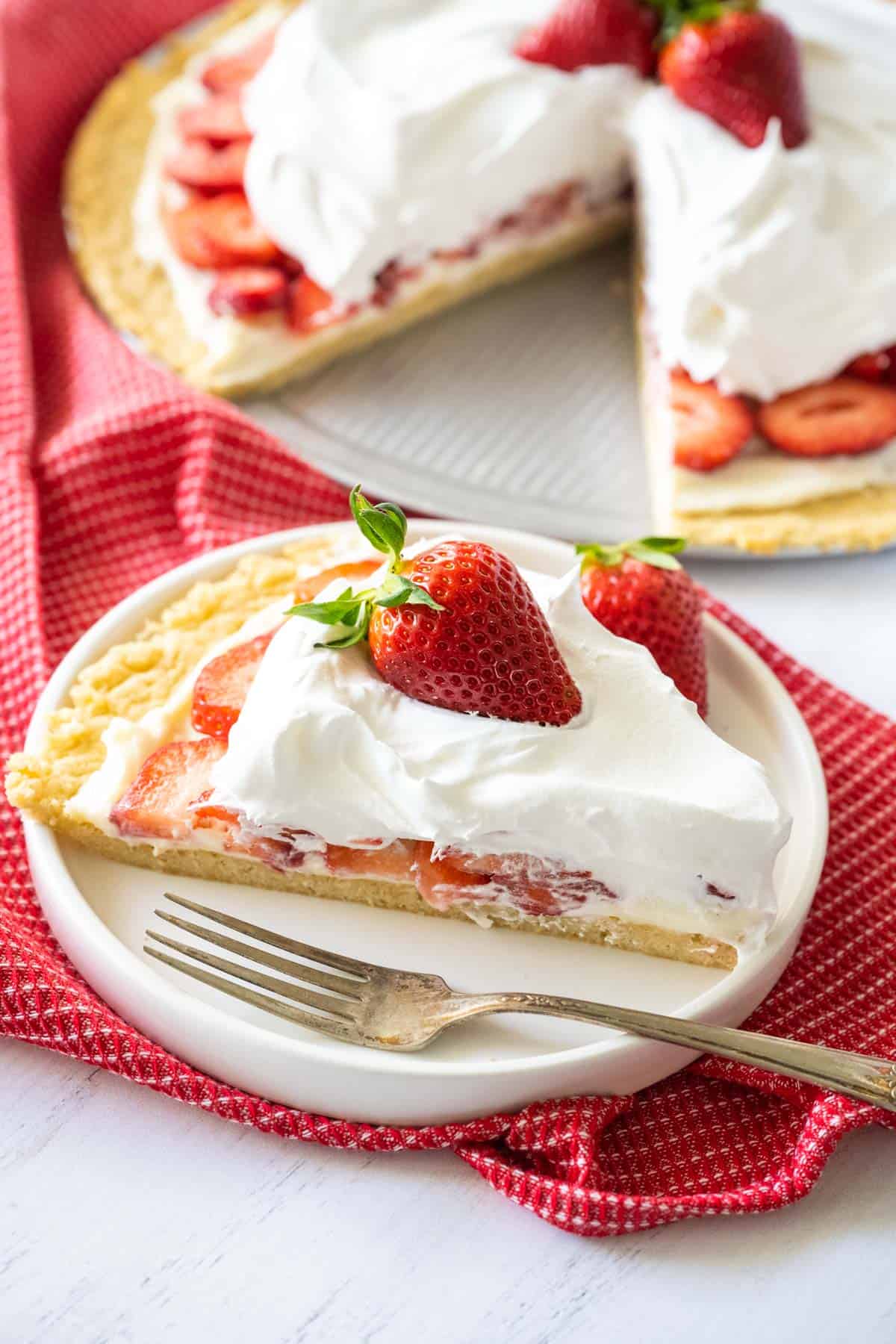Slice of strawberry dessert showing delicious layers of cookie crust, cheesecake filling, sliced strawberries, and whipped topping.