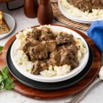 Beef tips over mashed potatoes on a white plate.