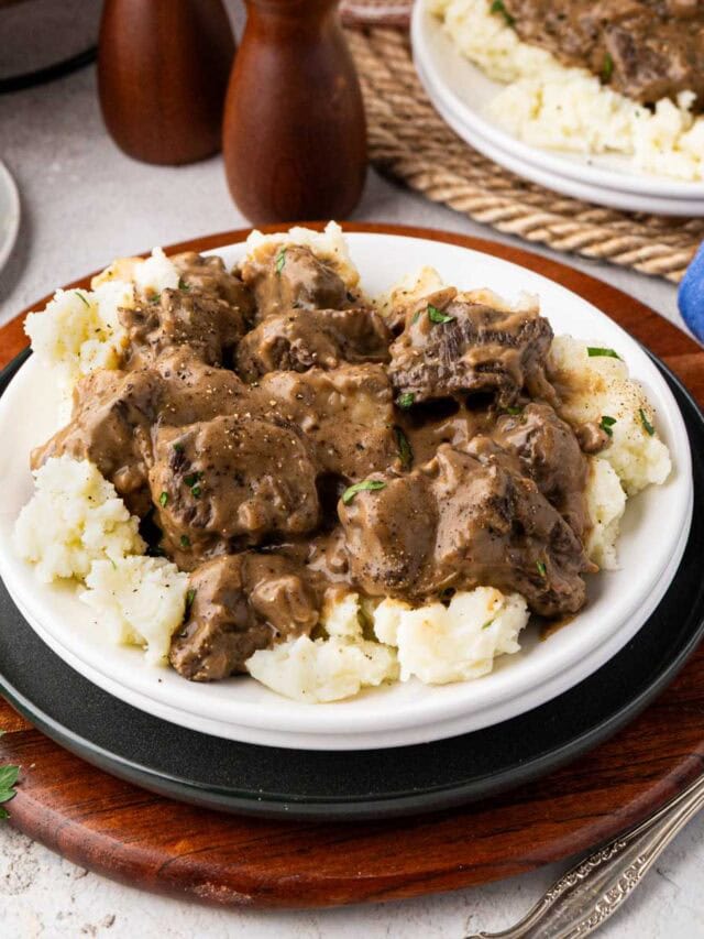Beef tips over mashed potatoes on a white plate.