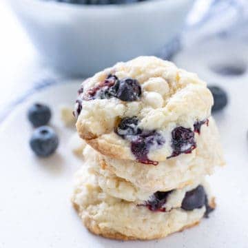Three blueberry cheesecake cookies stacked with blueberries scattered in the background.