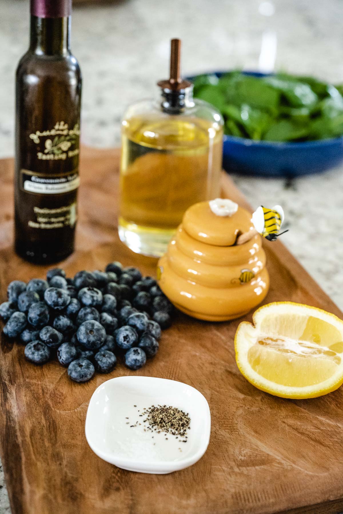 A wooden tray with balsamic vinegar, olive oil, blueberries, honey, lemon, and salt and pepper.