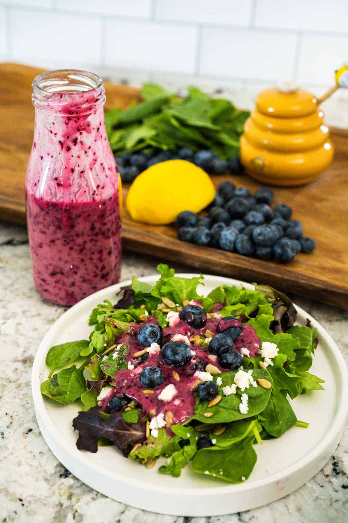 A spring blend of salad on a white plate with feta, blueberries and nuts topped with blueberry vinaigrette dressing. A bottle of the dressing and a wooden tray filled with blueberries, lemon, greens, and honey in the background.