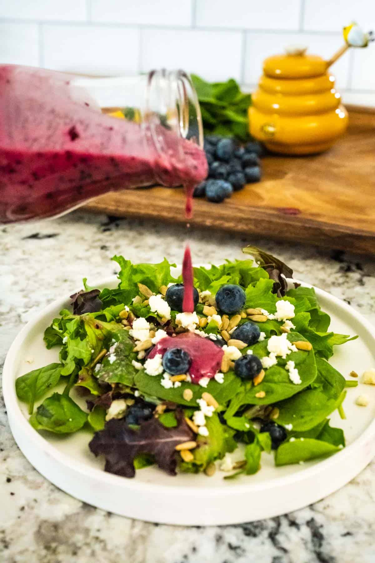 Pouring blueberry vinaigrette dressing onto a salad of fresh blueberries, feta, sunflower seeds, and spring greens.