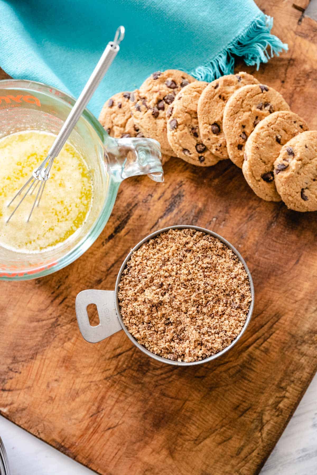 A wooden board with blue kitchen towel whole chocolate chips cookies and the ingredients needed to make crust - melted butter and chocolate chip cookie crumbs.