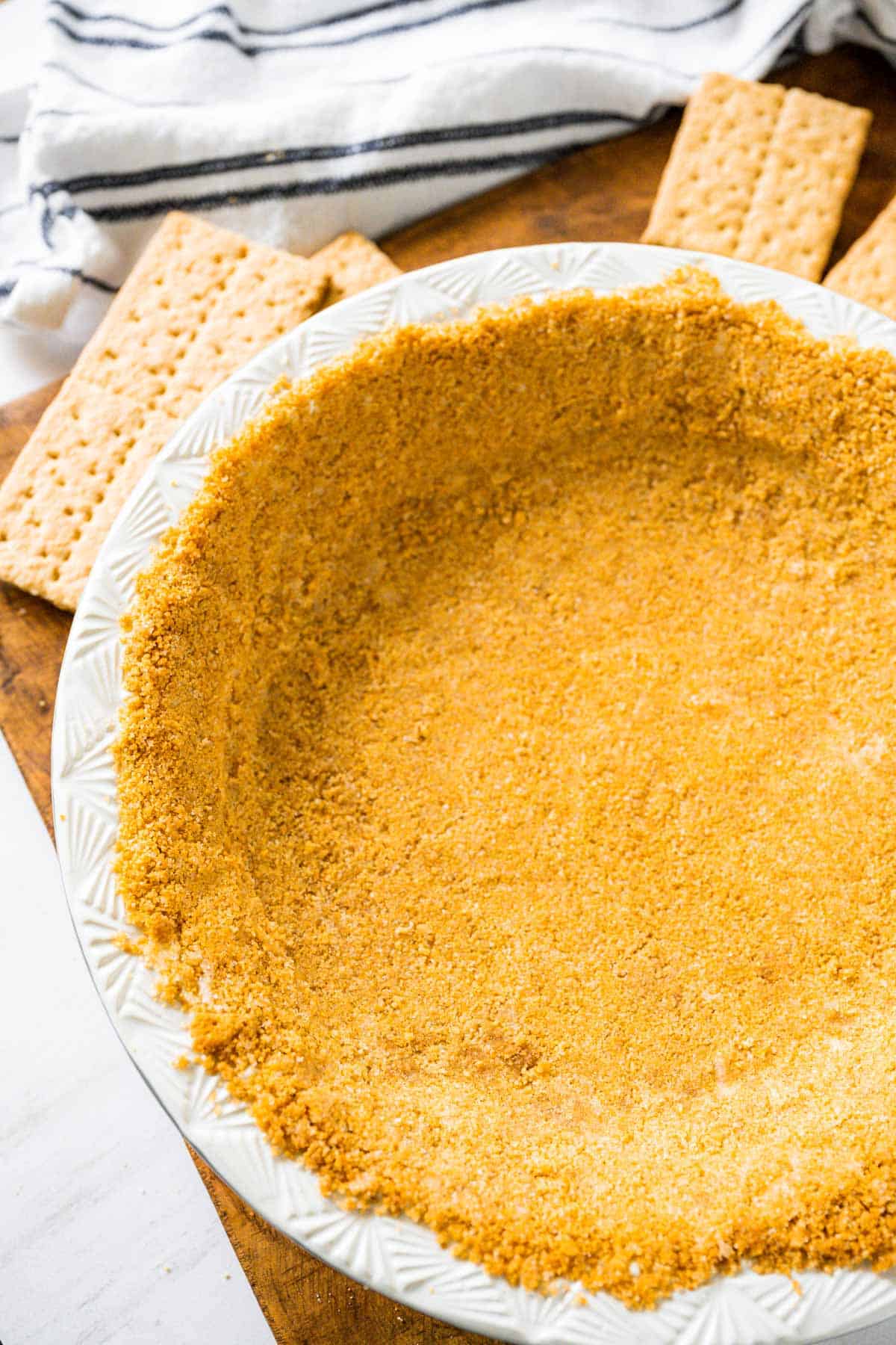 A homemade baked graham cracker crust in a white pie plate surrounded by graham crackers and a tea towel.