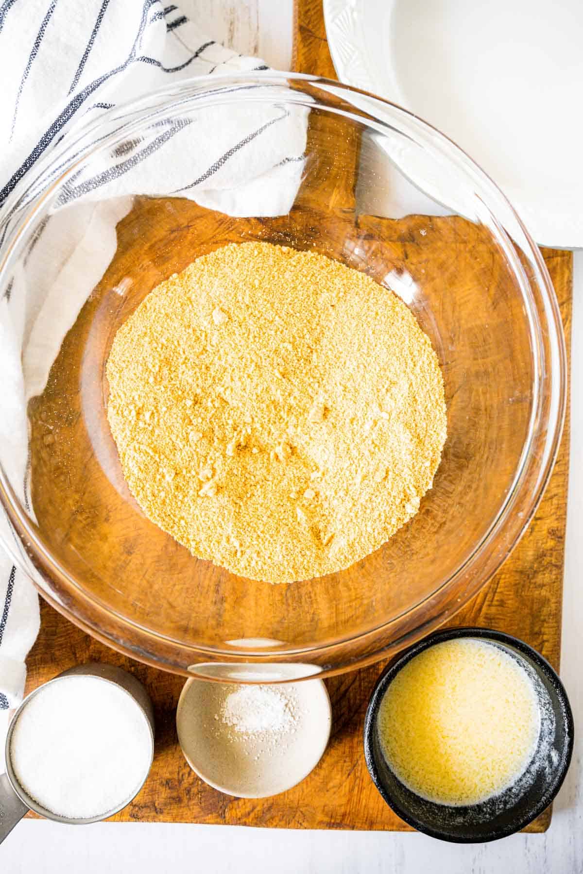 Crushed graham cracker crumbs in a bowl on a wooden board with sugar, salt, and melted butter.