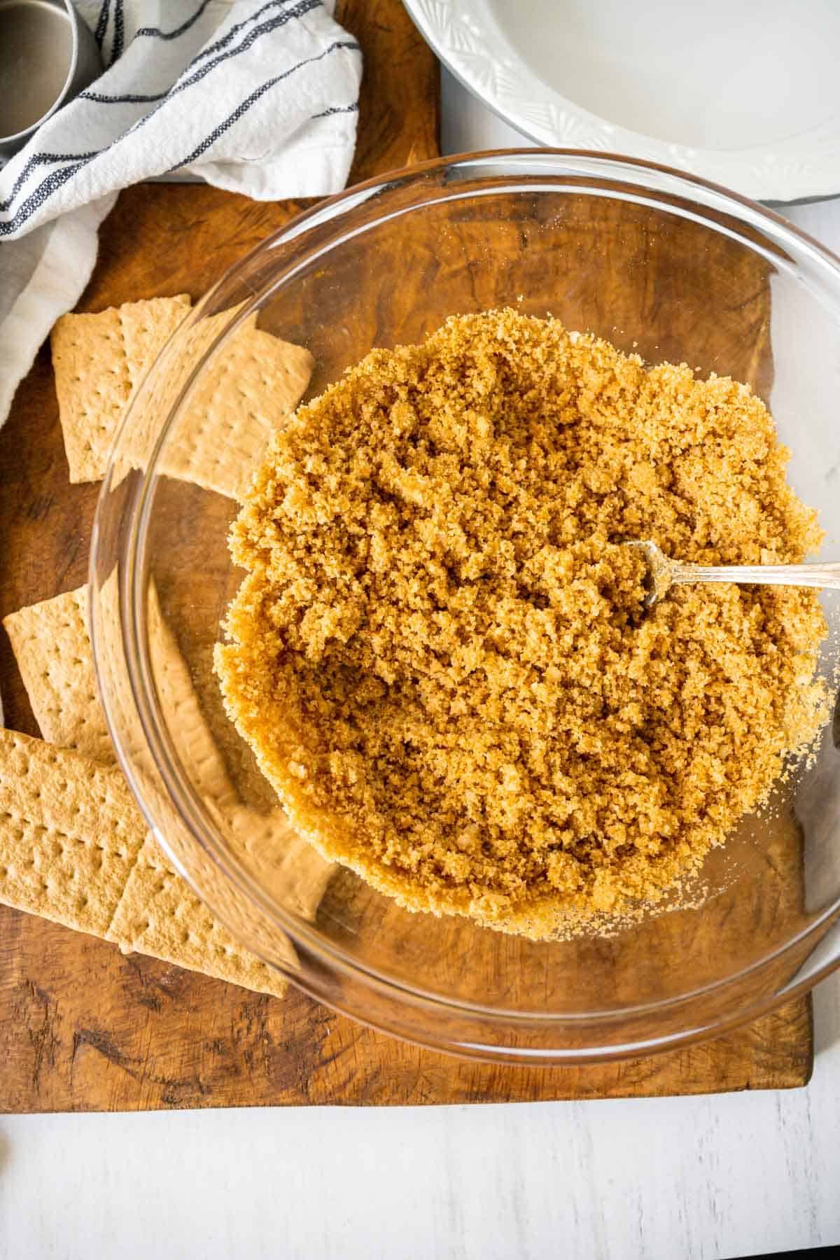 A bowl of graham crackers, sugar, butter and a pinch of salt mixed with a fork together on a wooden board with graham crackers scattered.