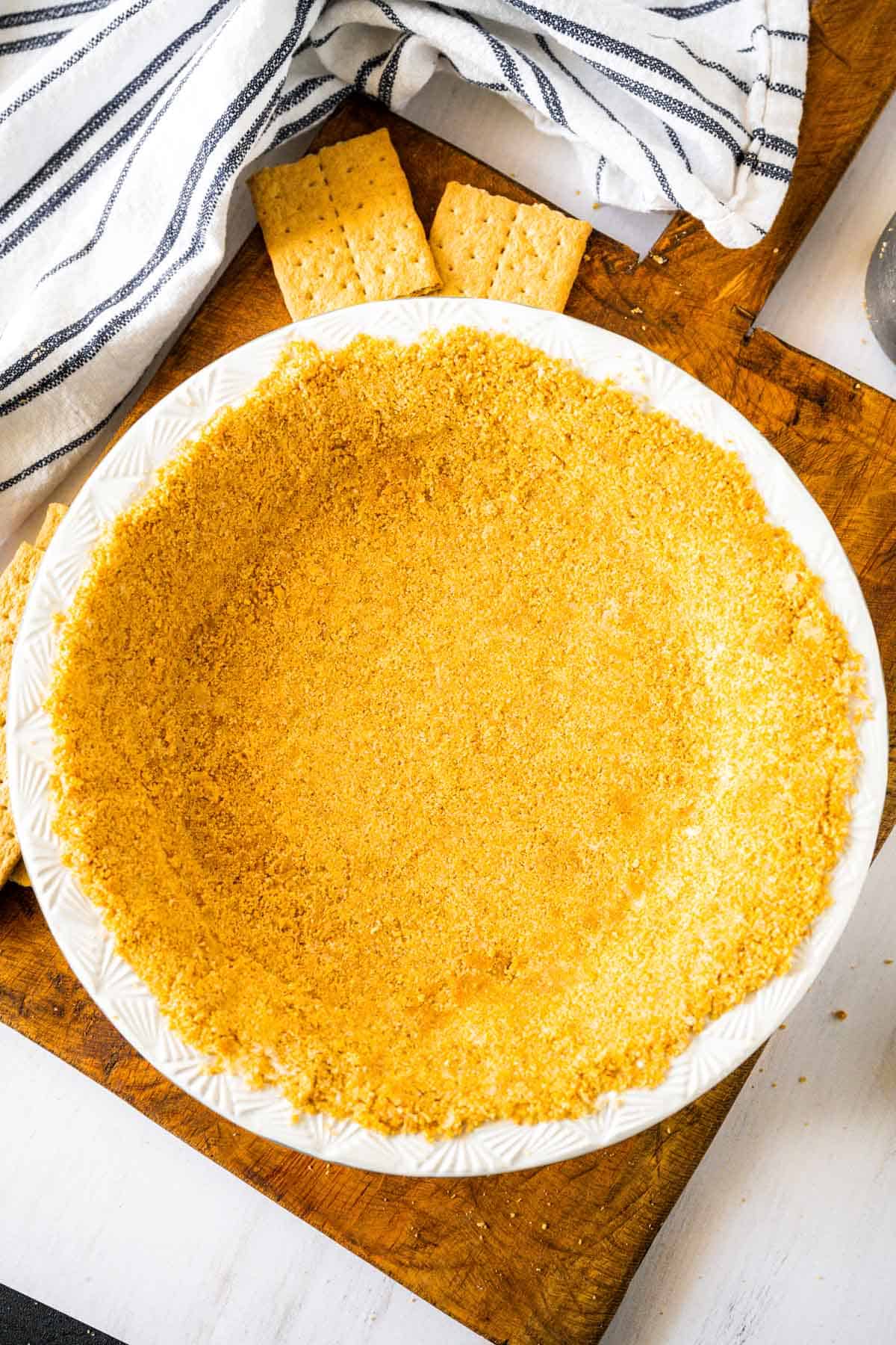 A no bake homemade graham cracker crust in a white pie plate cooling on a wooden board with scattered crumbs, crackers and a tea towel.