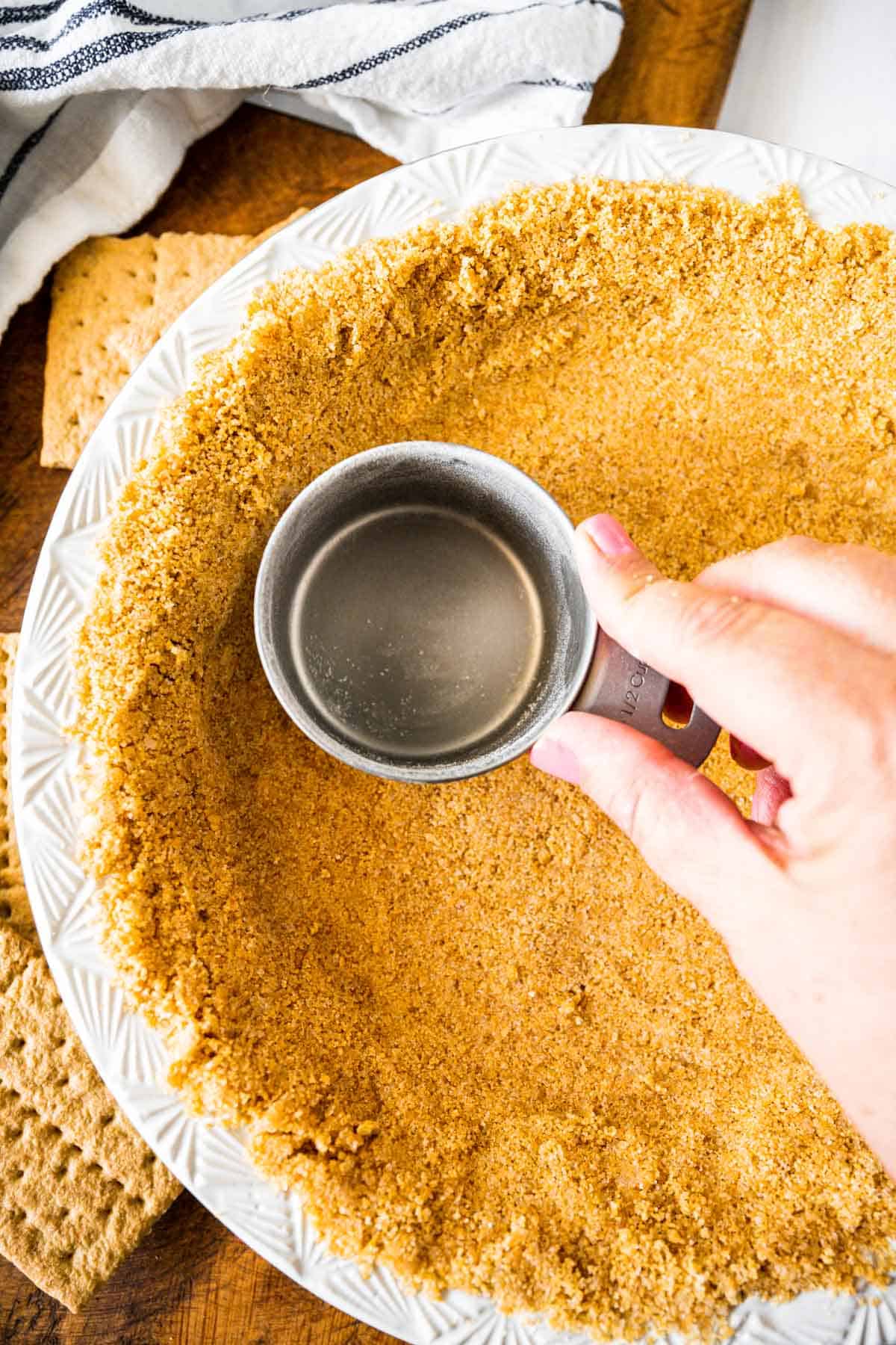 The hand of a person pressing a tin measuring cup into crumbs forming a homemade graham cracker crust in a white pie plate.