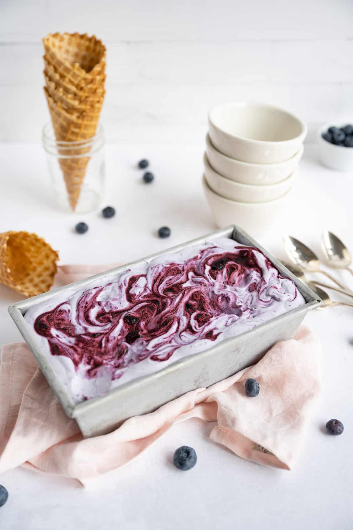 Container of blueberry ice cream set on a pink cloth with a stack of white bowls and a mason jar of ice cream cones in the backdrop.