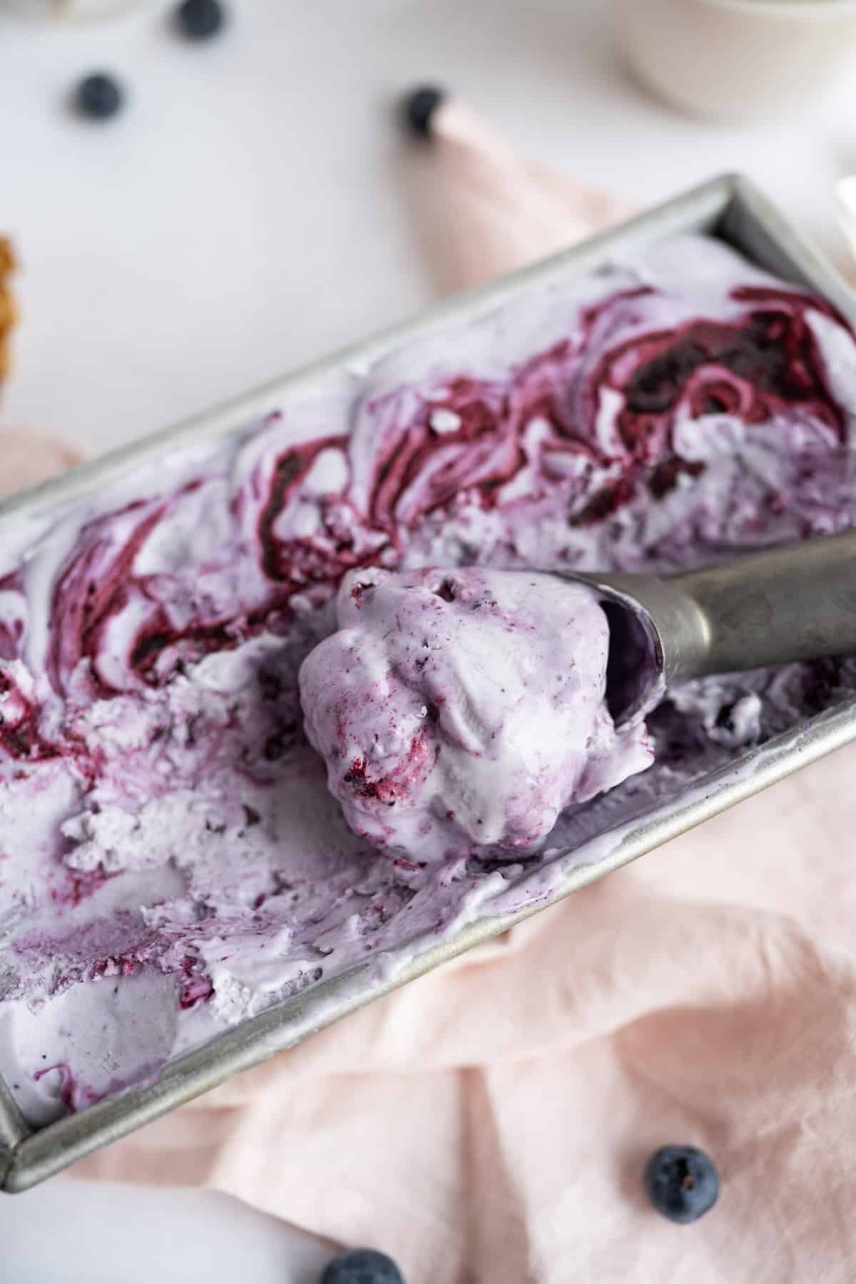 A metal loaf pan filled with no-churn blueberry ice cream being scooped with an ice-cream scoop.