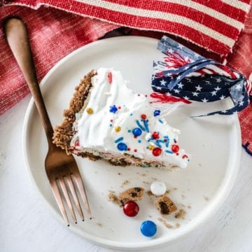 Overhead image of a slice of patriotic chocolate chip cookie candy cream pie on a white plate with cookie crumbles and m&m's on the plate along with a fork, with American flag decor in the background.