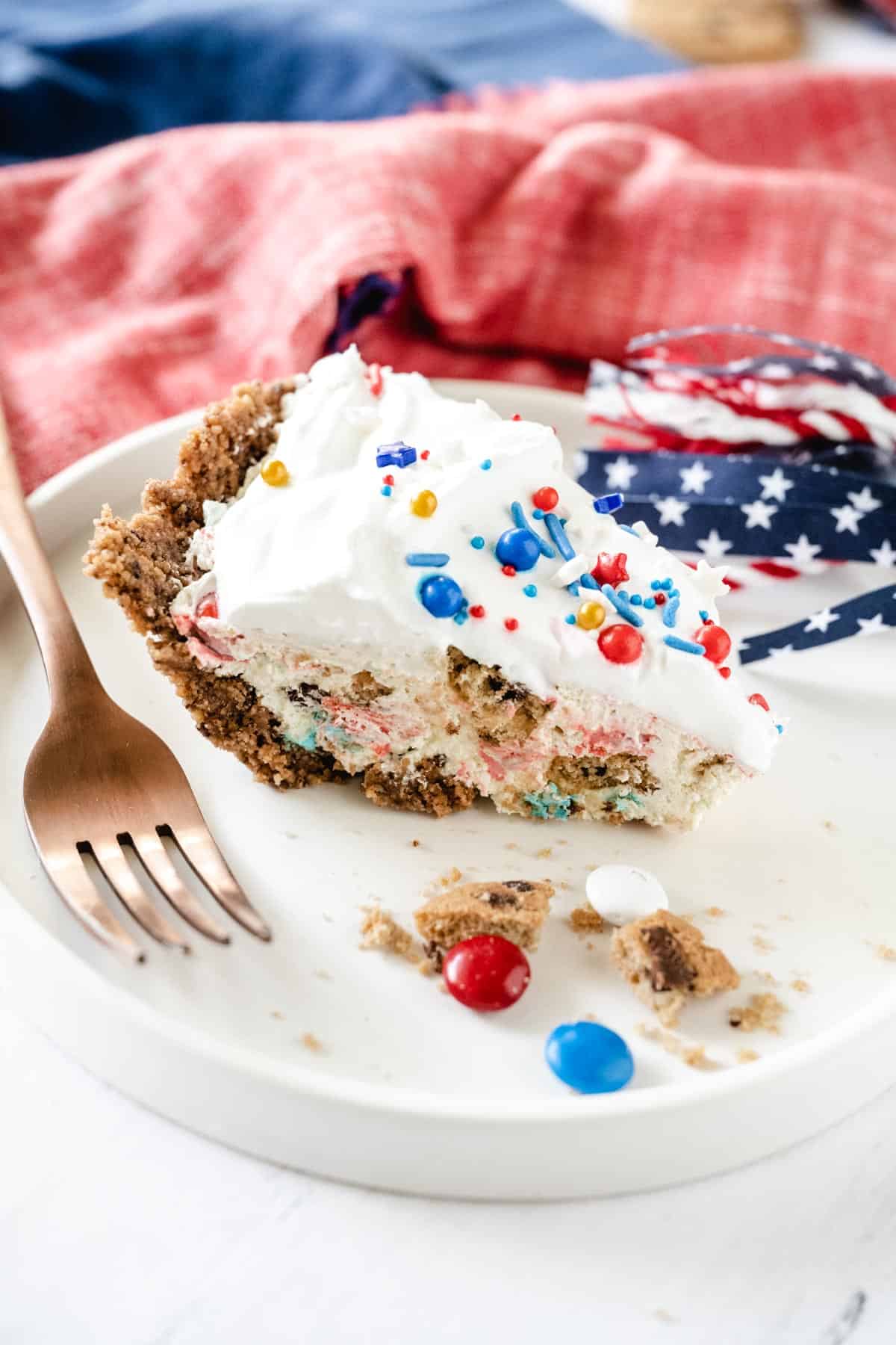 A slice of patriotic color themed slice of chocolate chip cookie candy cream pie on a white plate with cookie crumbs and red, white, and blue M&M's on the plate.