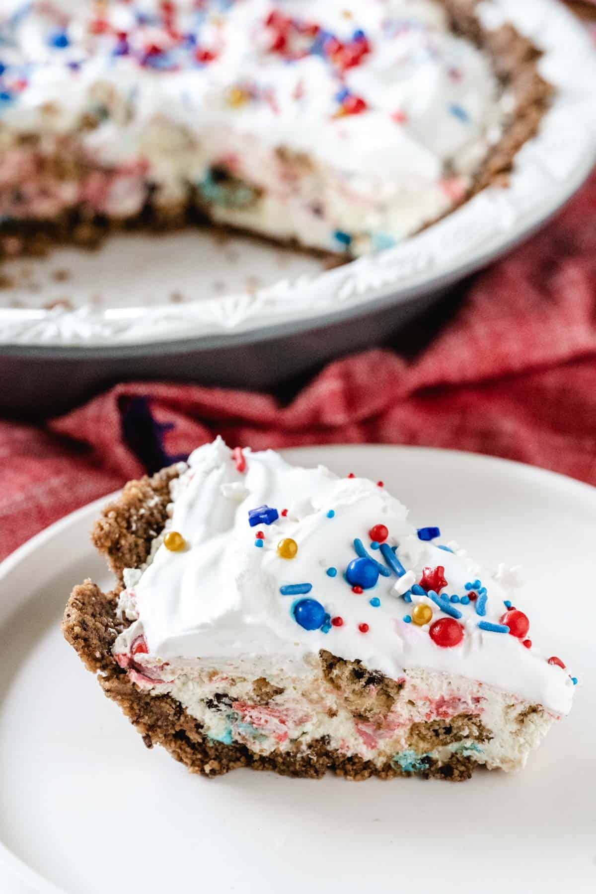 A slice of patriotic color themed slice of chocolate chip cookie candy cream pie on a white plate with the whole pie cut in the background.