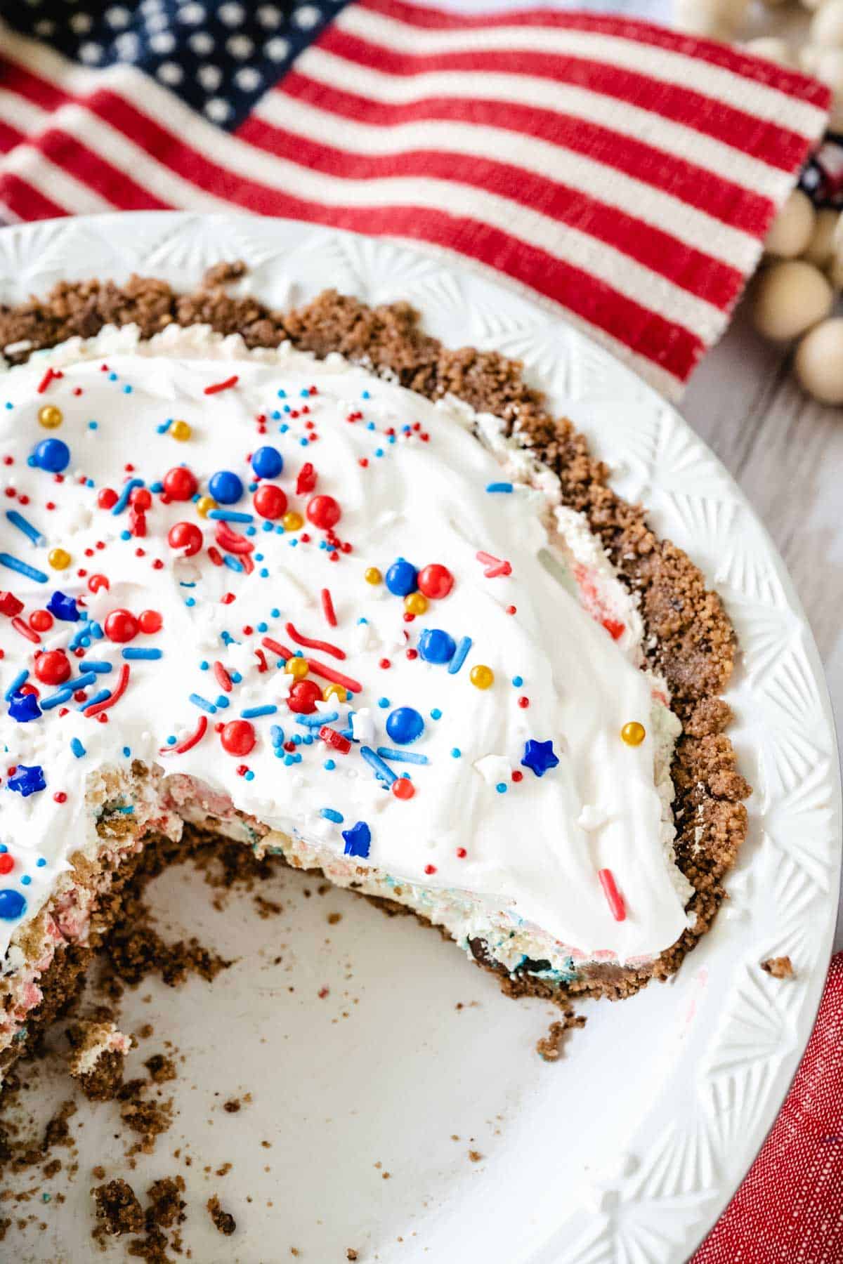 A whole chocolate chip cookie candy pie garnished with red, white, blue and gold sprinkles with slices removed in a white pie plate and an American flag in the background.