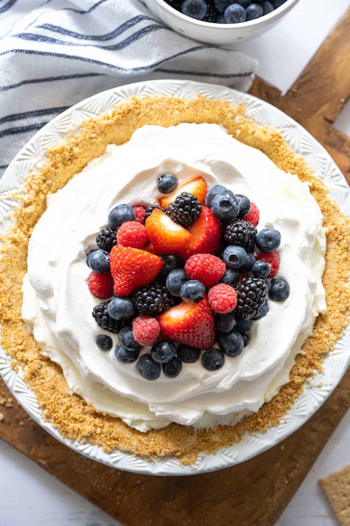 Overhead image of a whole red white and blueberry cream pie on a wooden board with scatter graham cracker crust crumbs and a bowl of berries.