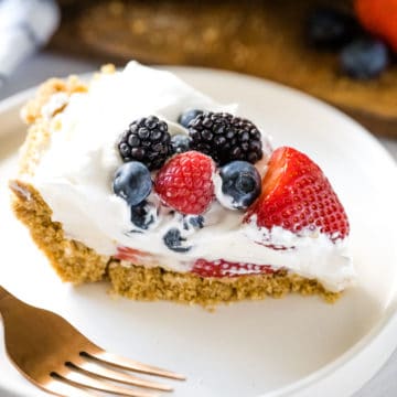 Image of a slice of red white and blueberry cream pie on a white plate with a gold fork shot up close showing all the layers in the pie.