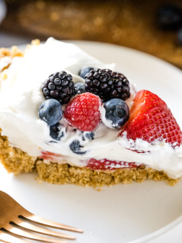 Image of a slice of red white and blueberry cream pie on a white plate with a gold fork shot up close showing all the layers in the pie.