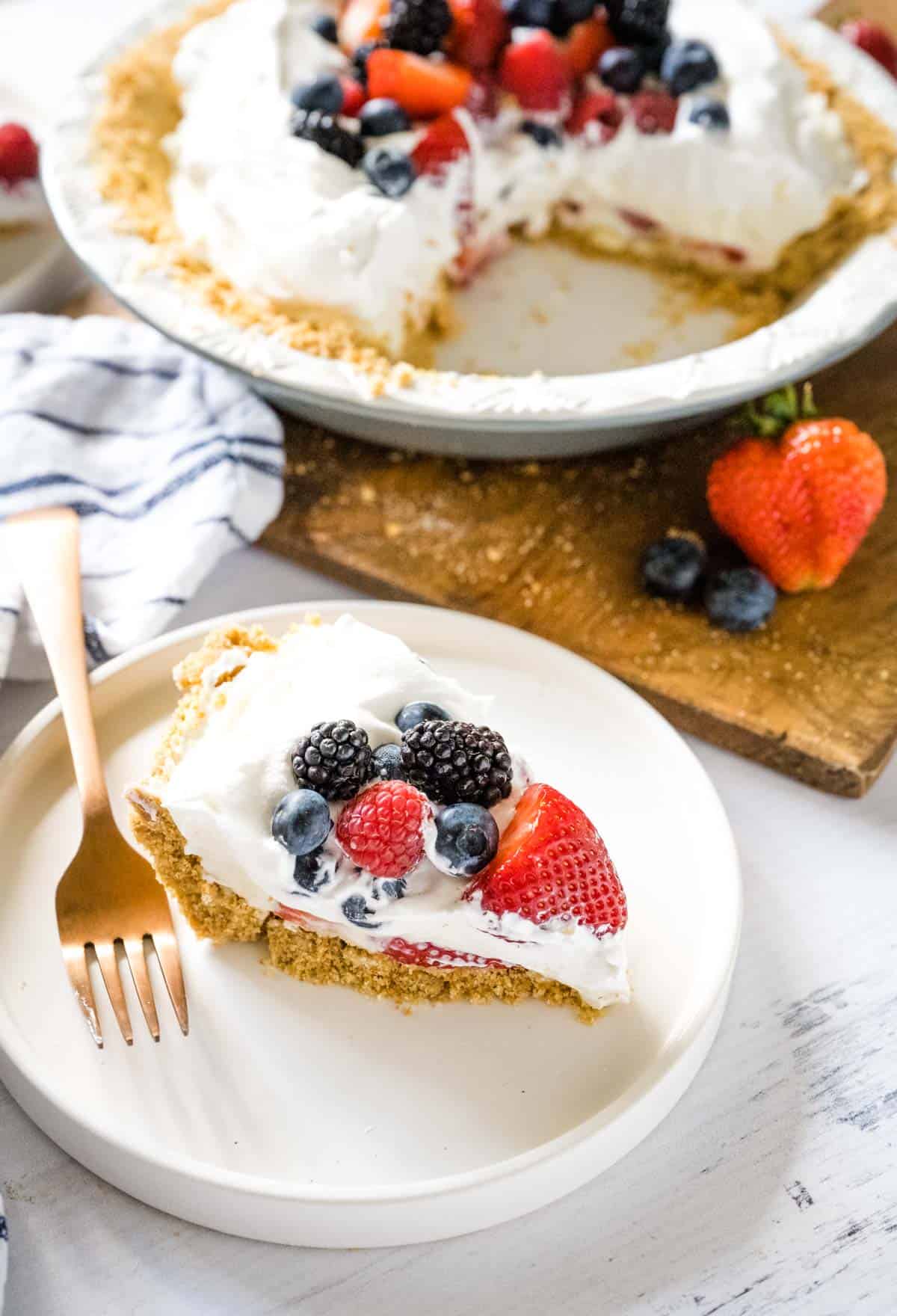 A slice of berry cream pie on a white plate set on a white wooden table with the whole pie cut into in the background.