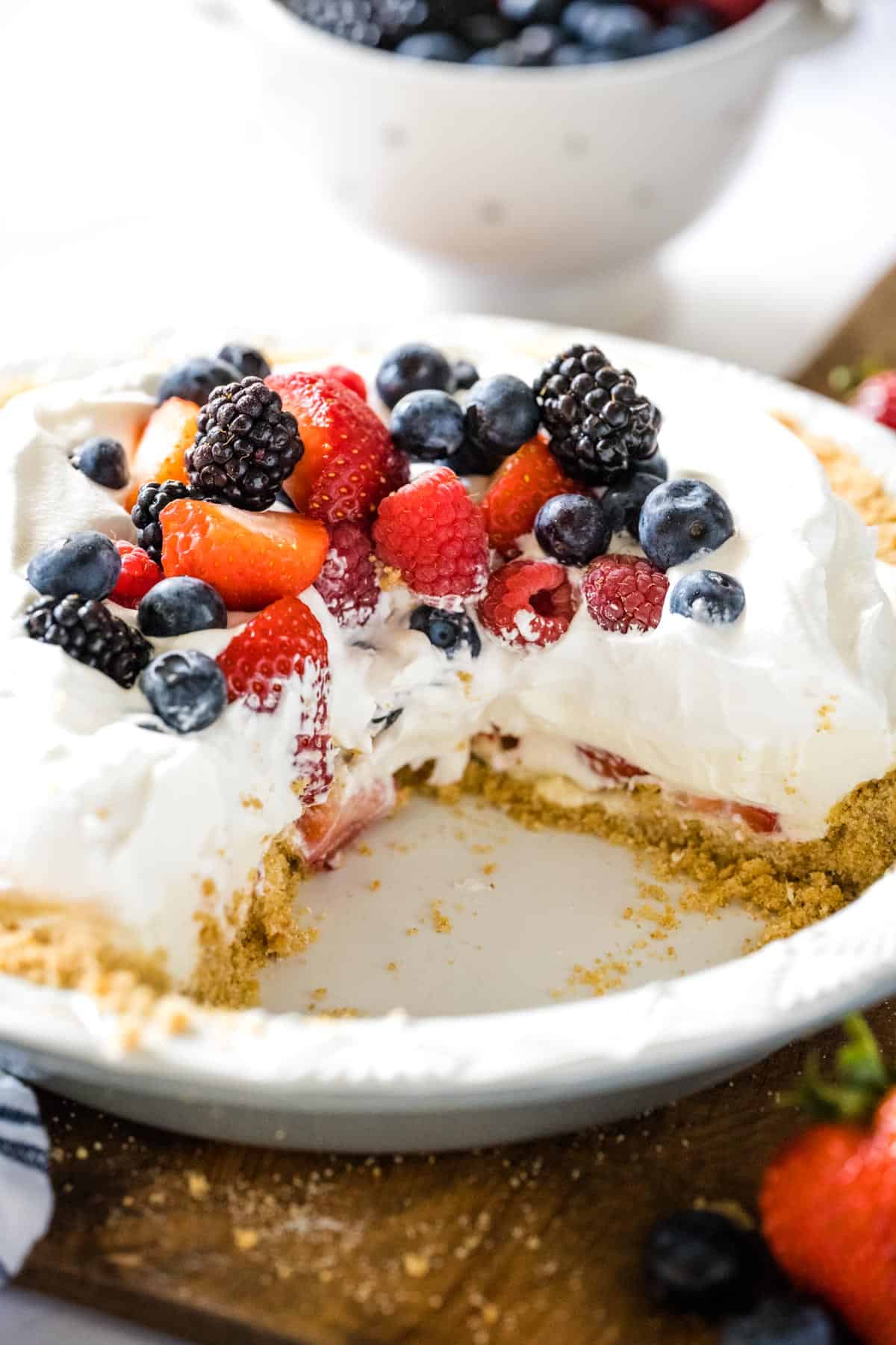 A white pie plate with a red white and blueberry cream pie that has been cut set on a wooden board with a strawberry and blueberries in the foreground.