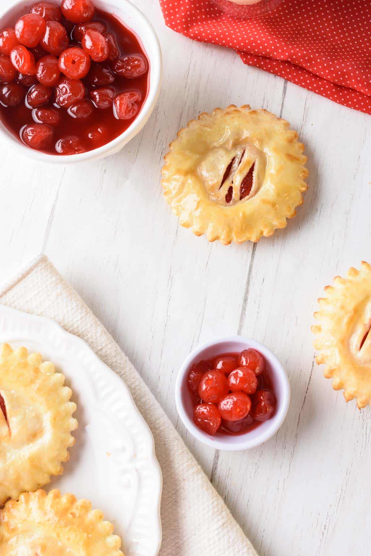 A white wooden table set with a plate of cherry hand pies, a bowls of cherry pie filling and a bright red napkin.