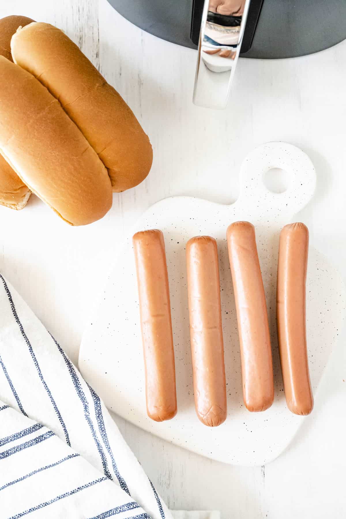 Image of ingredients needed for air fryer hot dogs - 4 hot dogs on a cutting board surrounded by hot dog buns, an air fryer, and a tea towel.