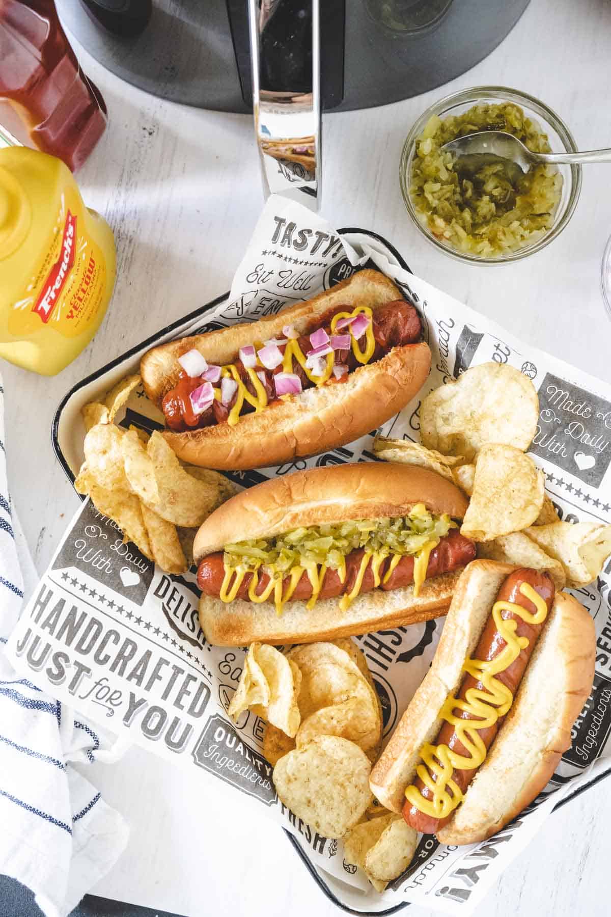 Three air fryer hot dogs in buns on a white tray surrounded by potato chips with pickled relish, mustard and ketchup set on the table.