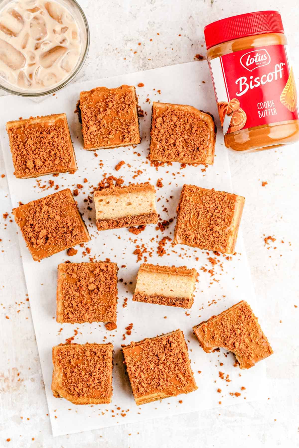 Slice Biscoff Cookie Butter Cheesecake Bars on parchment with a jar of Biscoff cookie butter and a glass of iced coffee in the background.