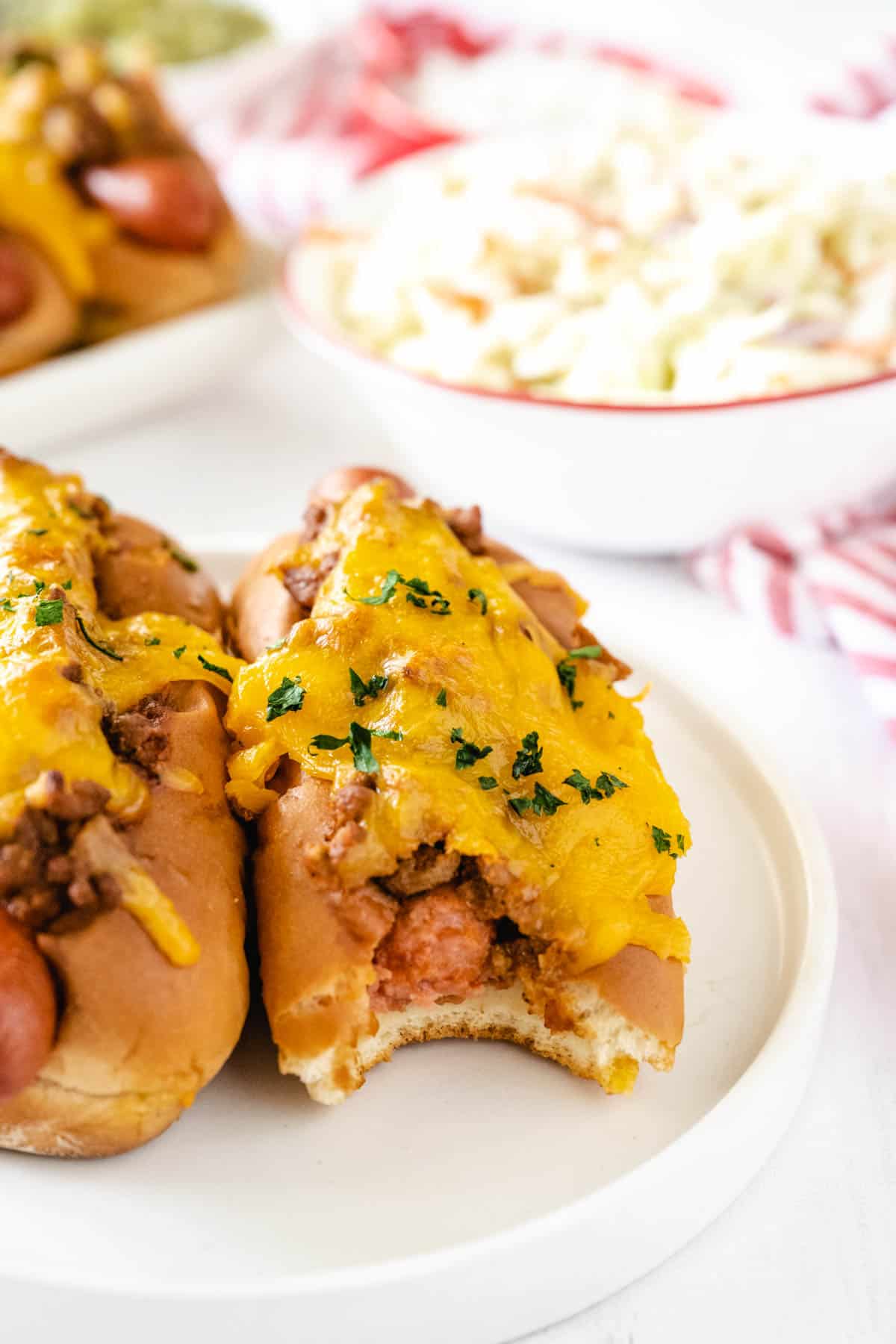 A hot dog topped with chili and cheese set on a white plate with a bite taken from the hot dog.