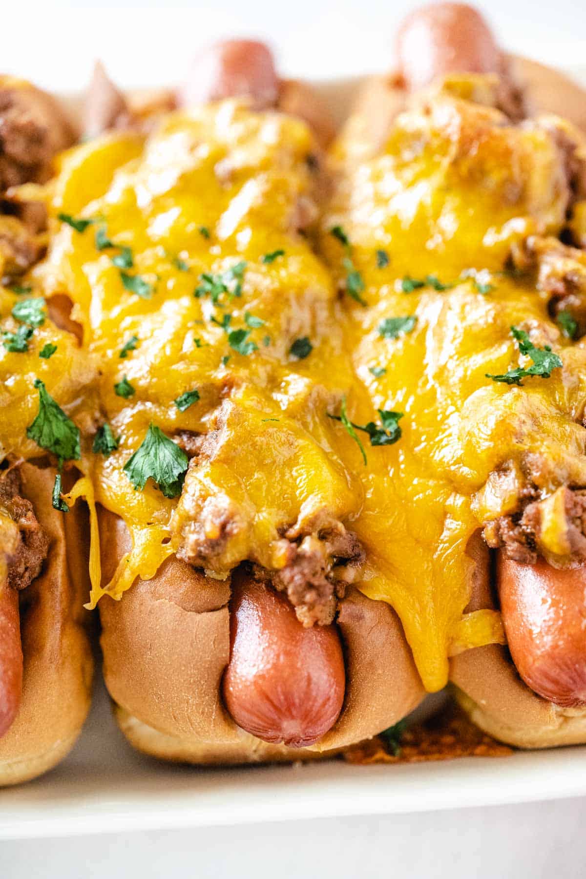An upclose image of showing glossy hot dogs smothered in chili and gooey melted cheddar cheese garnished with parsley.