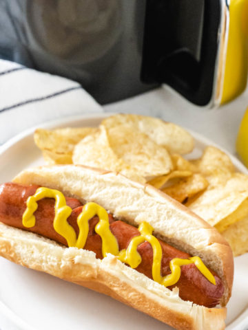 A hot dog cooked in the air fryer on a white plate along with potato chips with the air fryer in the background.