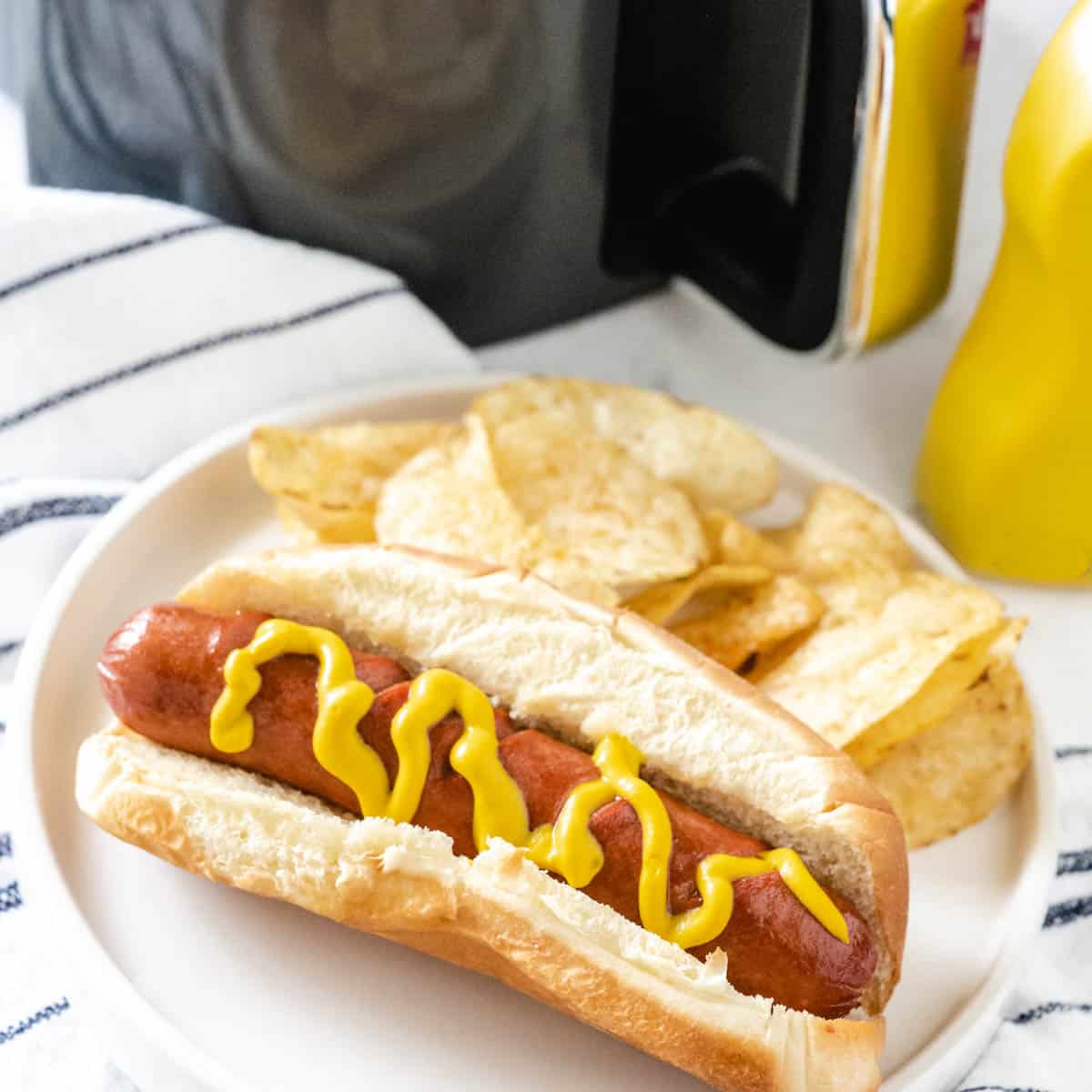 A hot dog cooked in the air fryer on a white plate along with potato chips with the air fryer in the background.