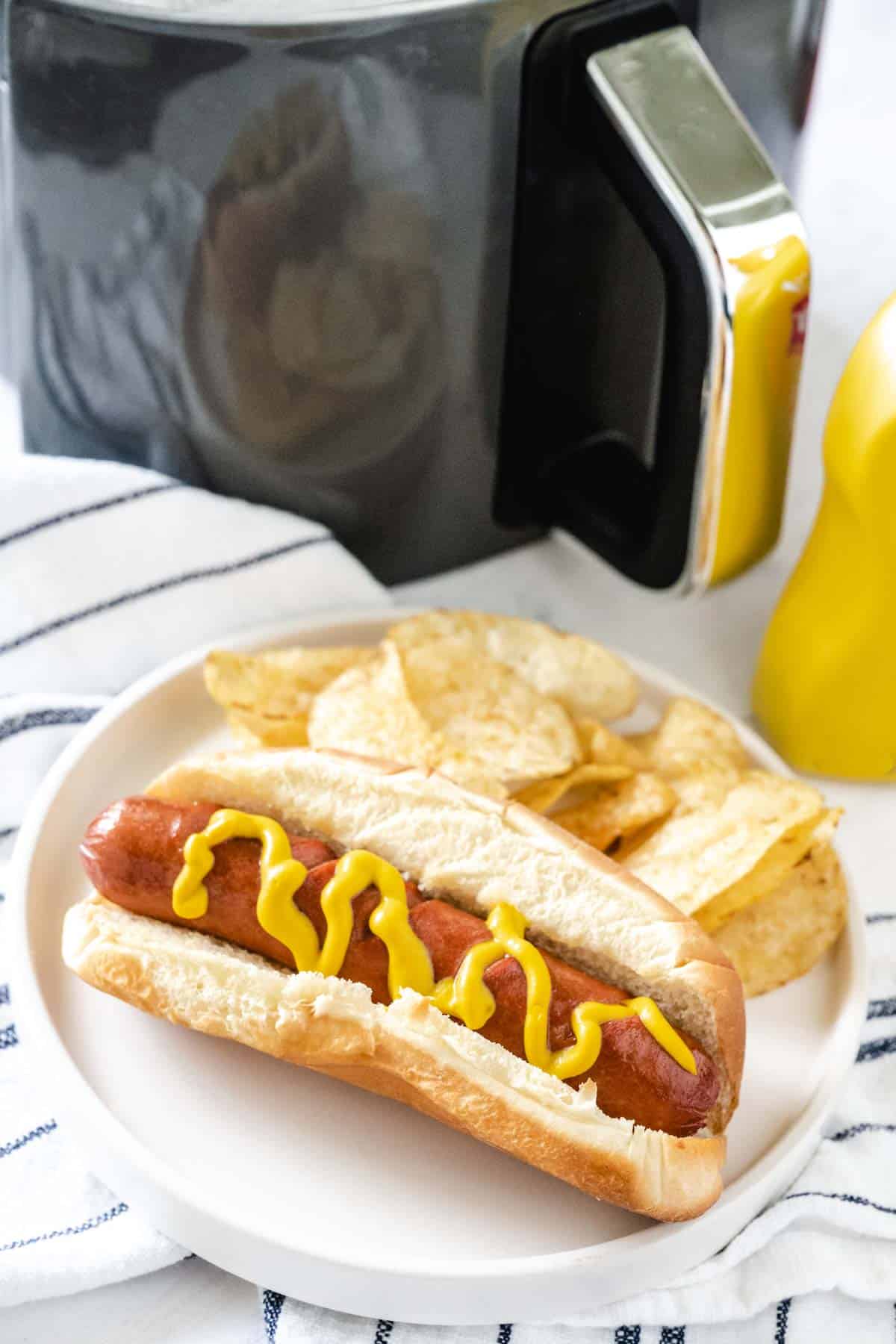 A hot dog cooked in the air fryer on a white plate along with potato chips with the air fryer in the background.