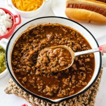 Hot dog chili sauce in a white enamel pan with a black rim being scooped out with a ladle.