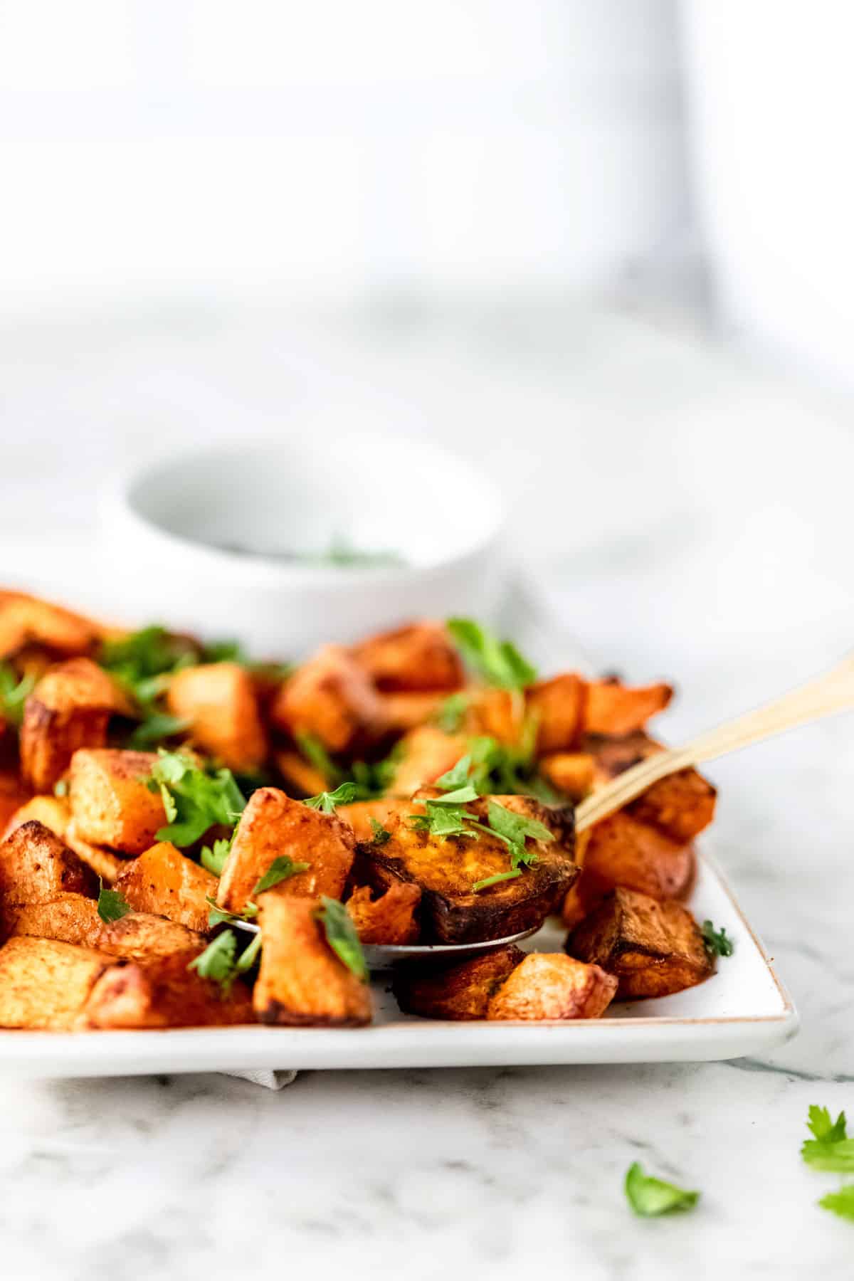 A side view image of sweet potatoes being lifted off of a white plate with a gold spoon.