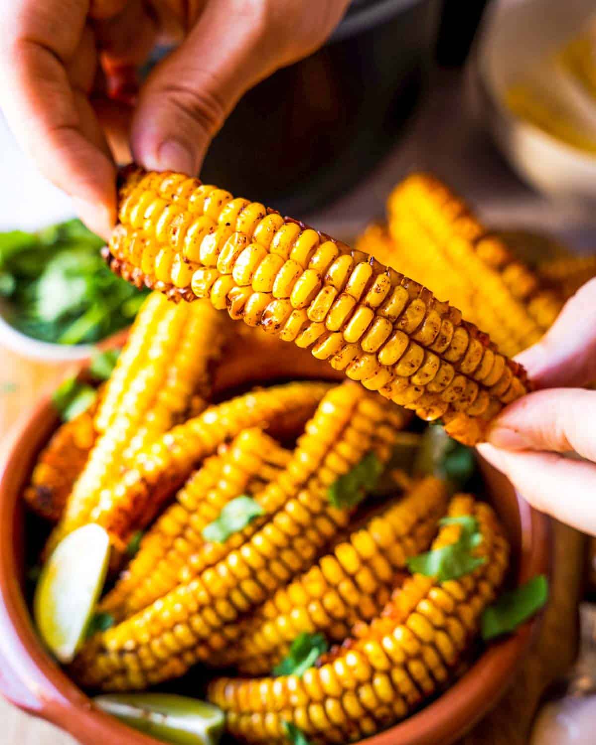 Hand holding a corn riblett with a bowl full of corn ribs in the background.