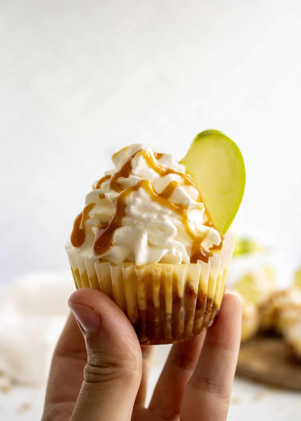 A hand holding up a mini caramel apple cheesecake in a cupcake paper topped with whipped cream, an apple slice, and drizzled with caramel sauce.