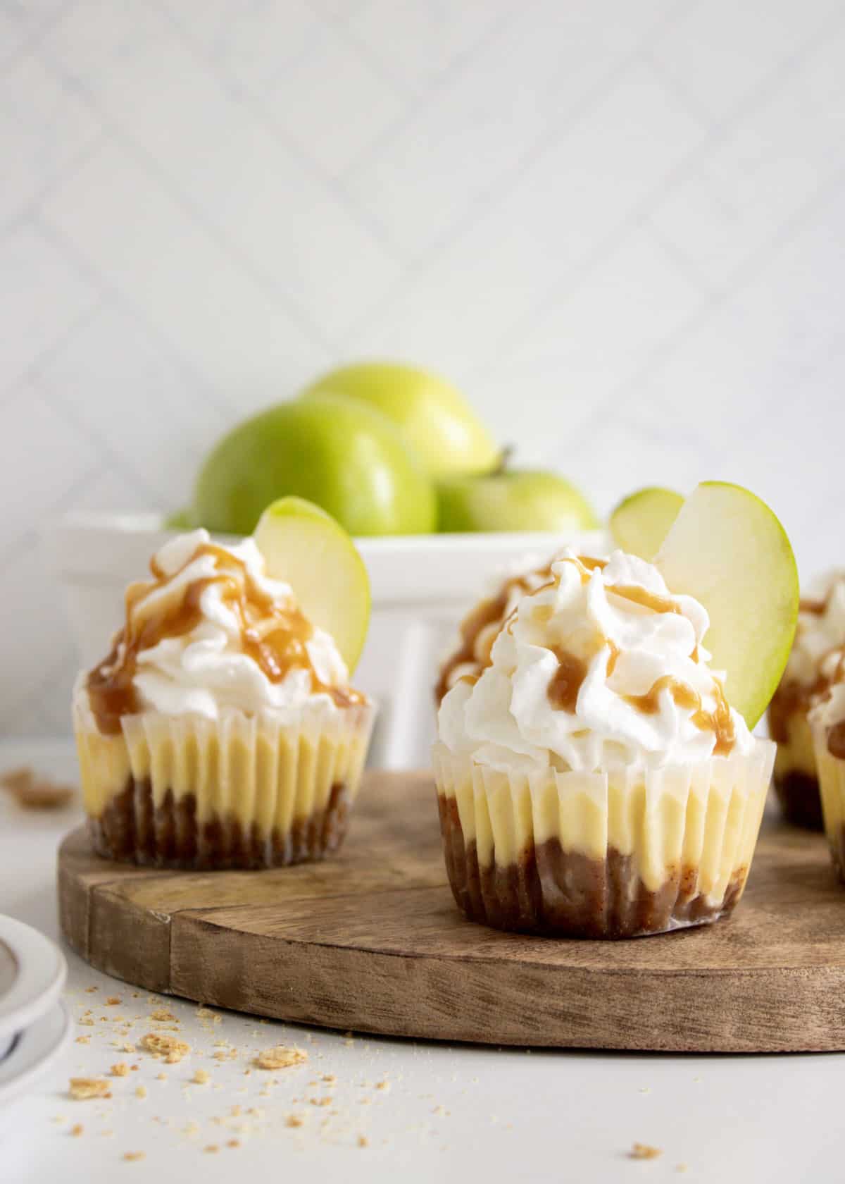 A wooden tray of caramel apple cheesecakes with a bowl of green apples in the background.