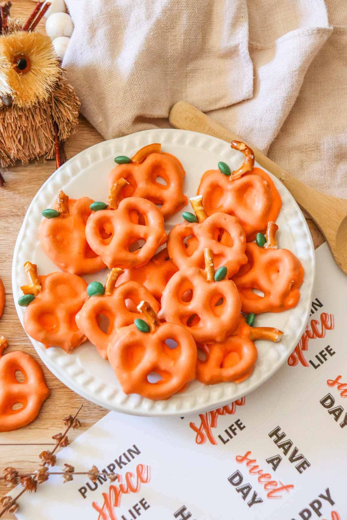 A white plate filled with chocolate covered pretzel pumpkins set on a table with a wooden spoon and a cream tea towel.