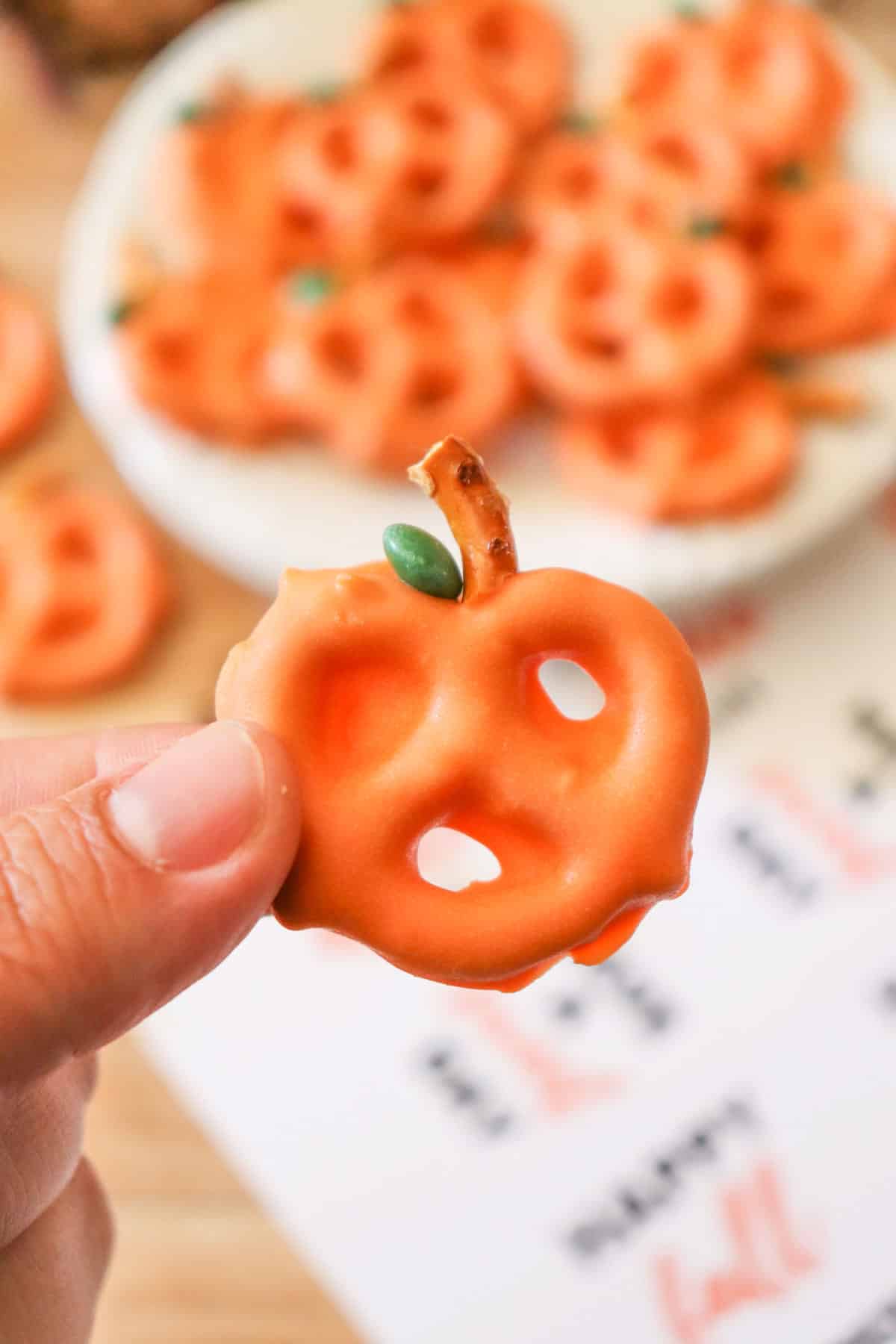 Someone holding a candy coated pumpkin pretzel so that it can be seen up close.