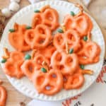 A white plate full of pretzels covered in orange colored chocolate decorated to look like a pumpkin.