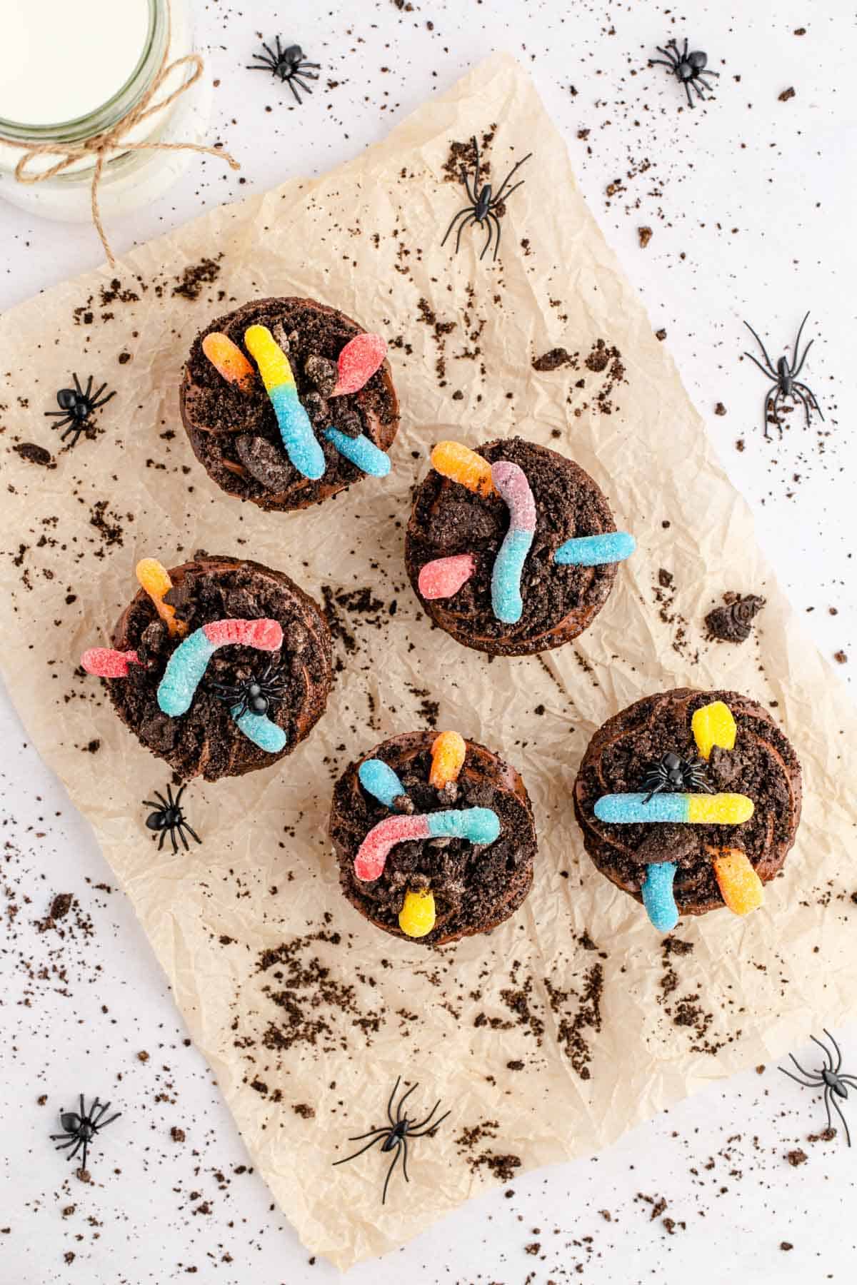 overhead image of a batch of gummy worm dirt cupcakes set on parchment paper with scatter cookie crumbs on a white table.