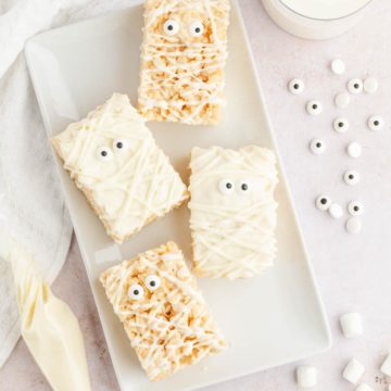 An overhead image of a white plate filled with mummy rice krispies with a piping bag of white chocolate, candy eyeballs, and mini marshmallows scattered around the plate.