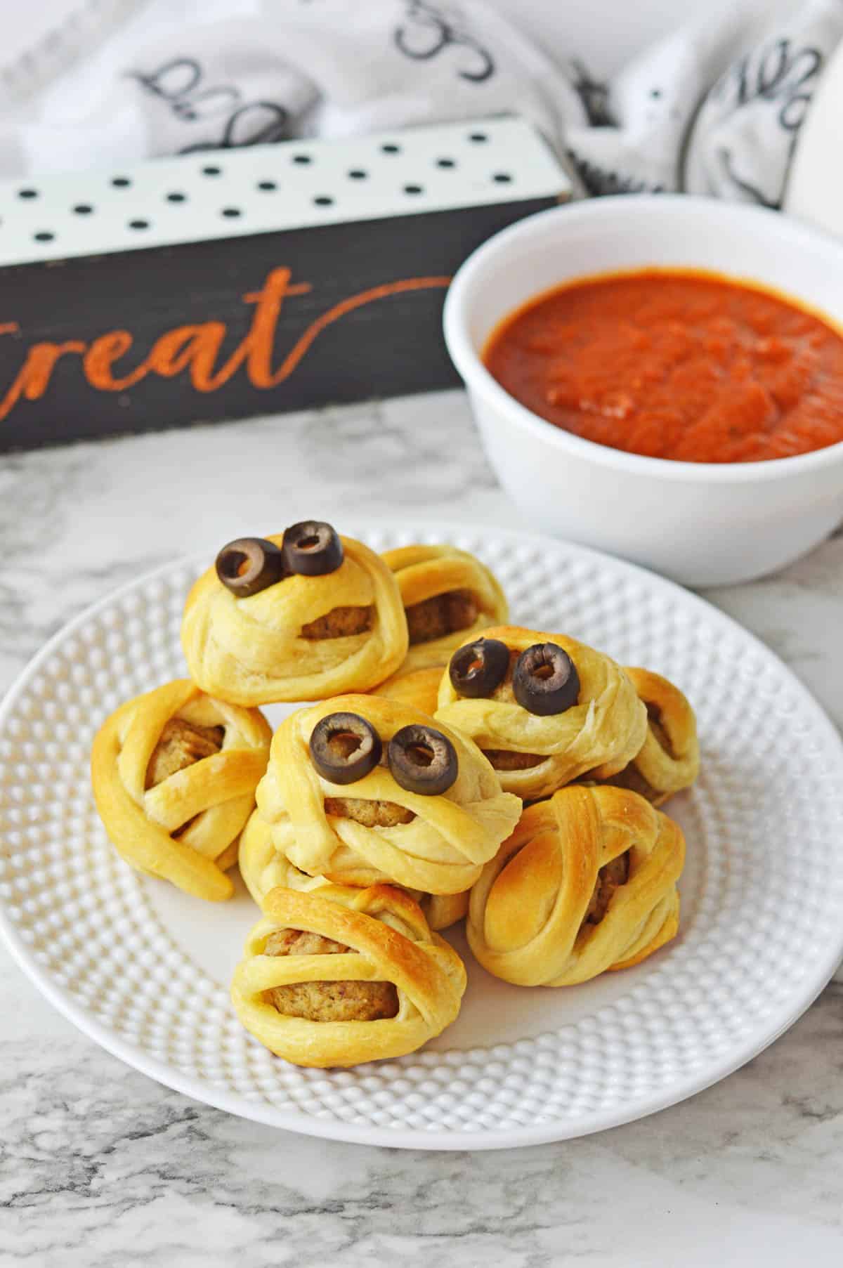 A white plate full of mummy meatballs with a bowl of marinara sauce in the background.