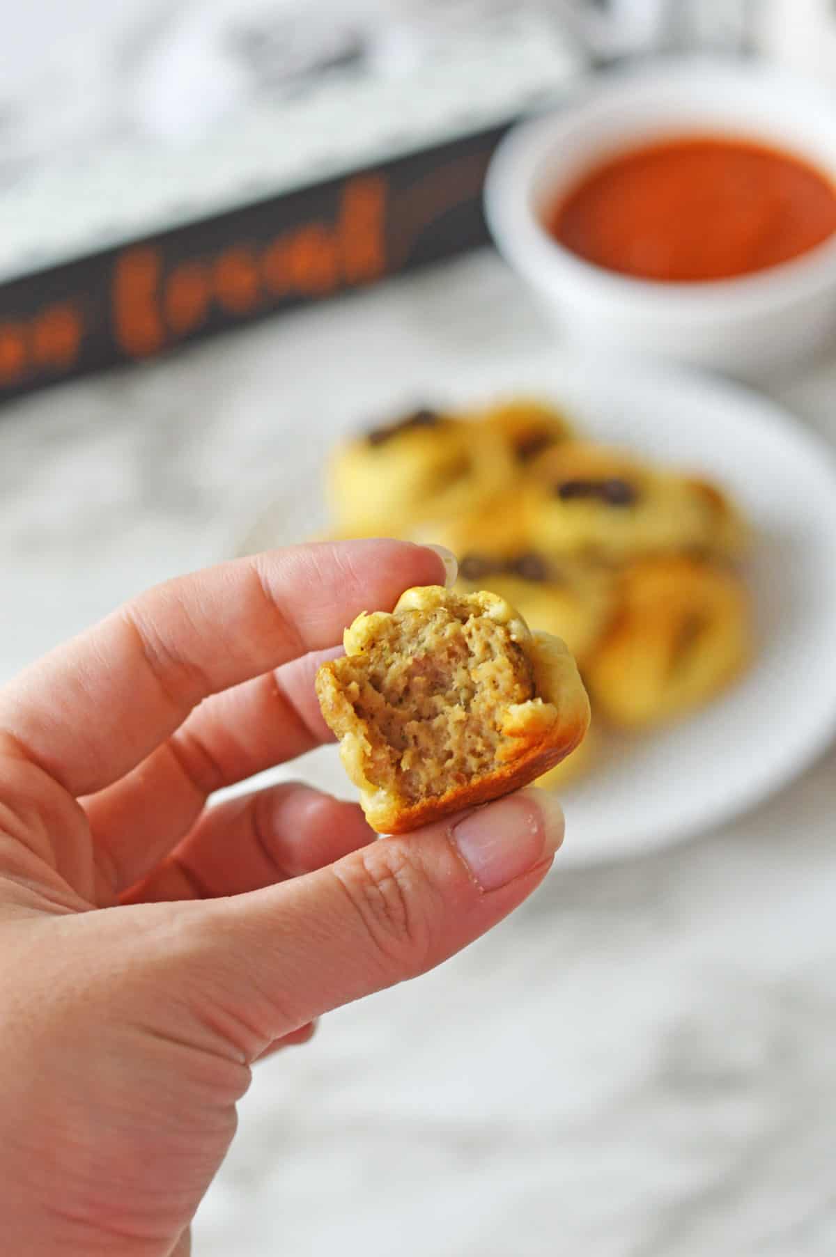 A hand holding up a mummy meatball with a bite taken out to show the center of the appetizer.