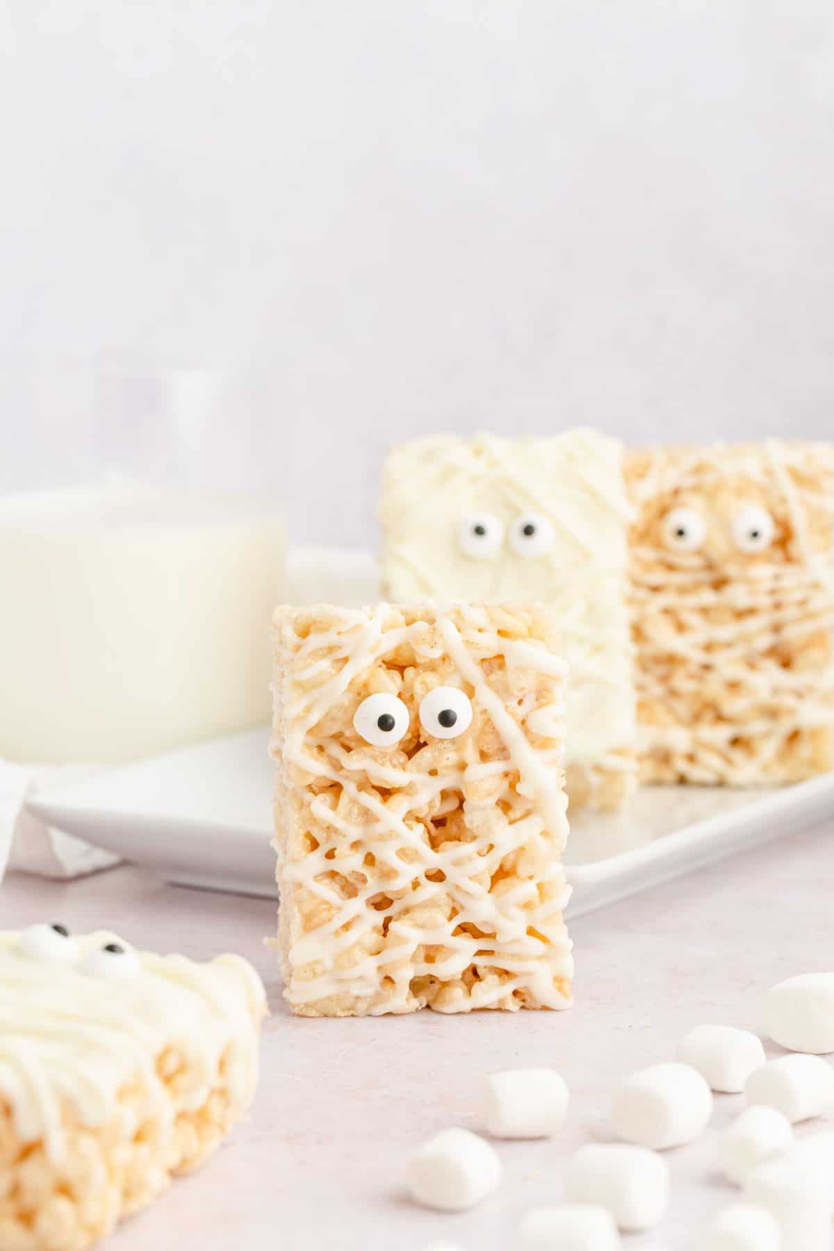 Rice krispie treats decorated like mummies with white chocolate and candy eyes propped up with a glass of milk and marshmallows.