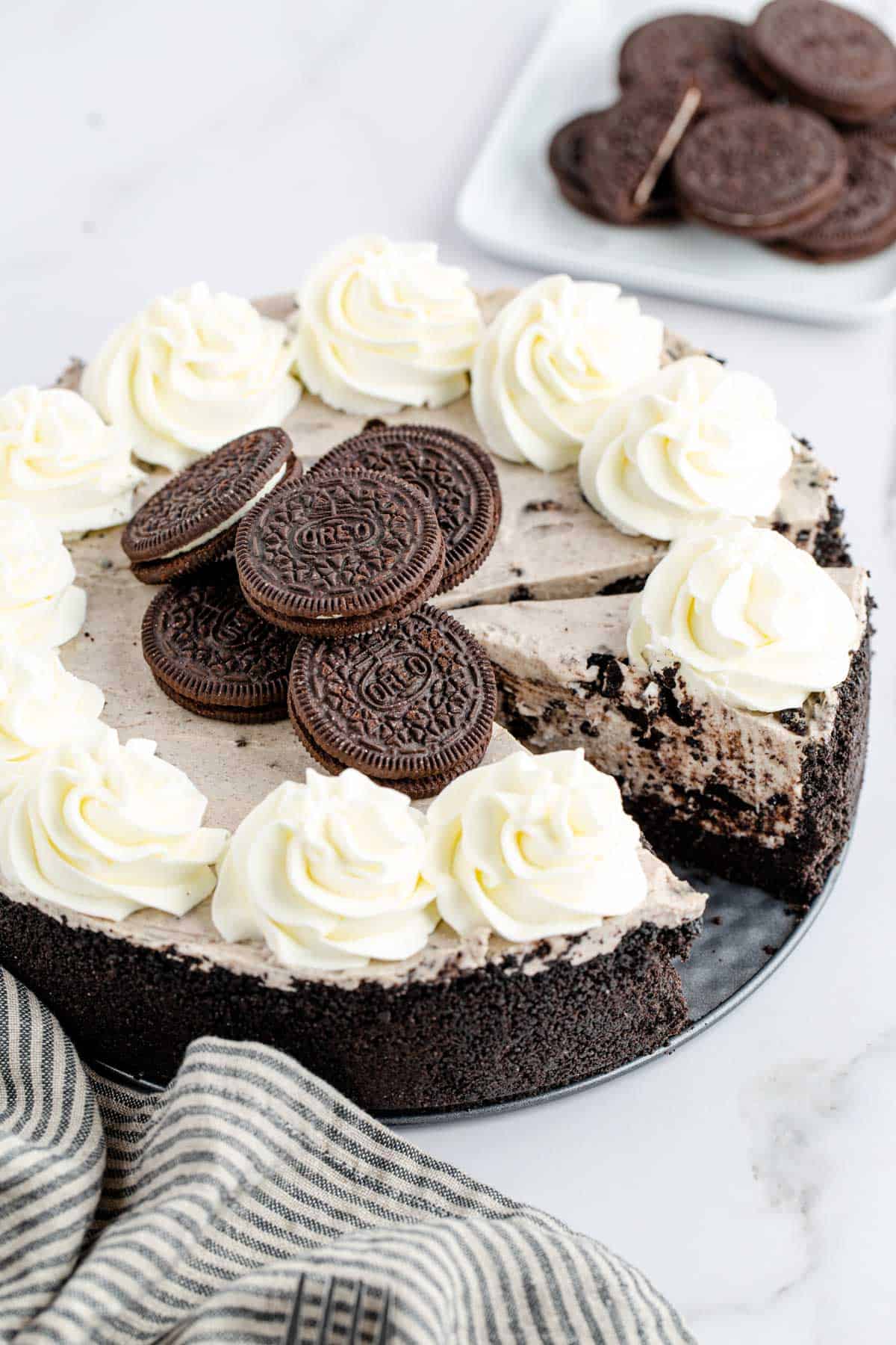 A partially sliced oreo cheesecake set on a marble table with a black striped tea towel set to the side and a bowl of Oreos in the background.