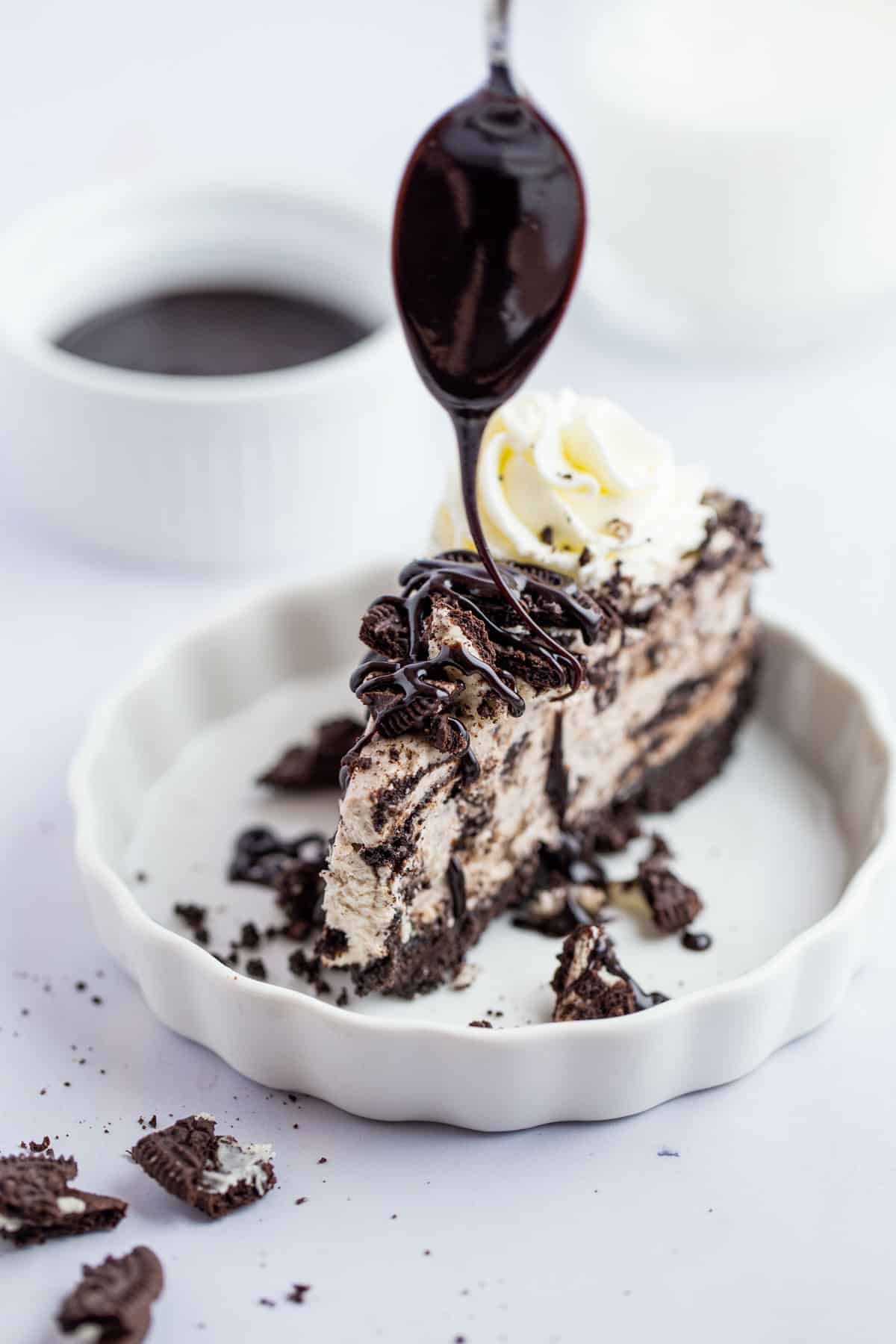 An action shot of chocolate syrup being spooned over the top of a slice of oreo cheesecake.