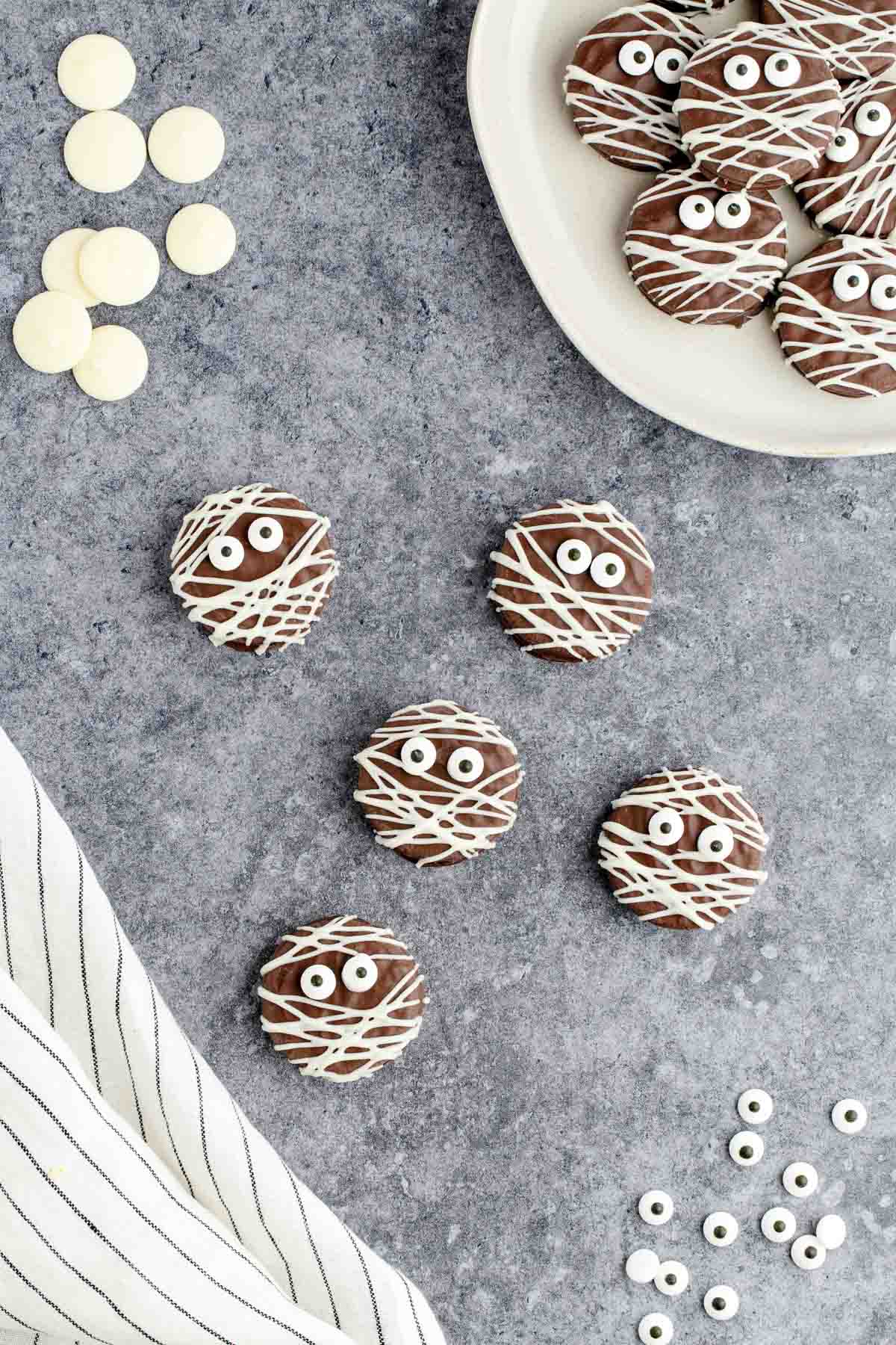 A grey table with fudge Oreo mummy cookies set with candy eyeballs, white chocolate melts, a black and white striped tea towel and a plate full of mummy cookies.