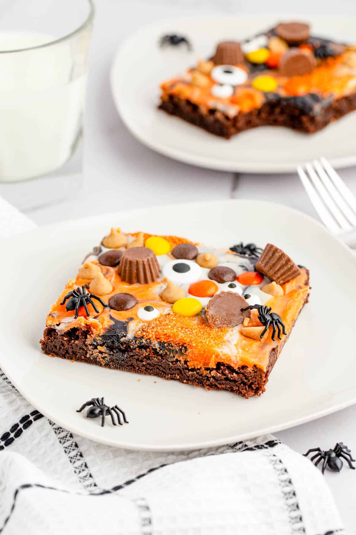 A slice of Peanut Butter Halloween Eyeball Brownies on a white plate with a white tea towel trimmed in black and scattered plastic spiders.